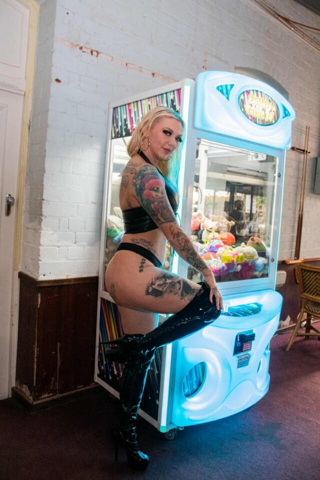 Tattooed skimpy barmaid Tiffy Texas poses confidently in a black leather-look bikini and thigh-high boots beside a glowing claw machine in a country pub.