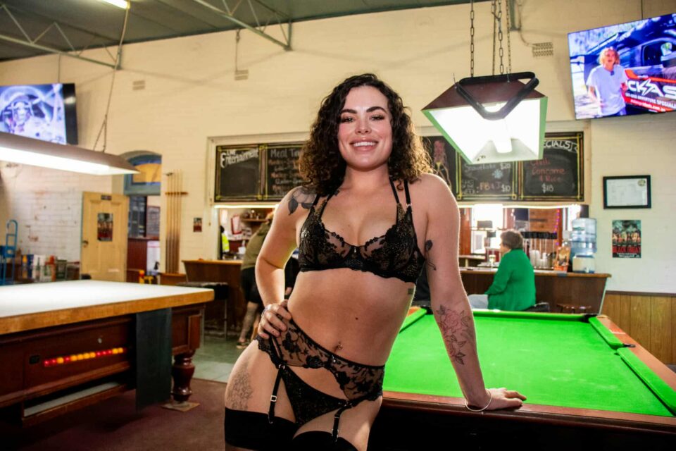 Bailey Weston, a skimpy barmaid in black lingerie, smiling confidently while leaning on a pool table at a WA country pub.