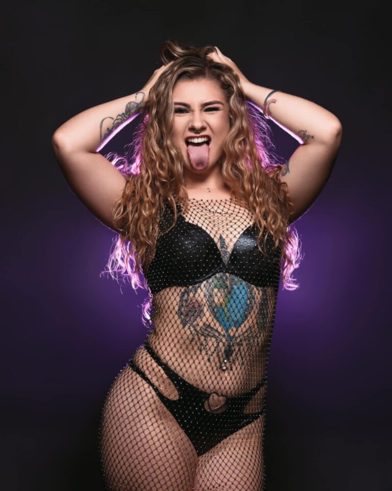 Zeena Zee, a confident and expressive skimpy barmaid, poses in black lingerie and mesh with her tongue out, showing off her tattoos under dramatic lighting.