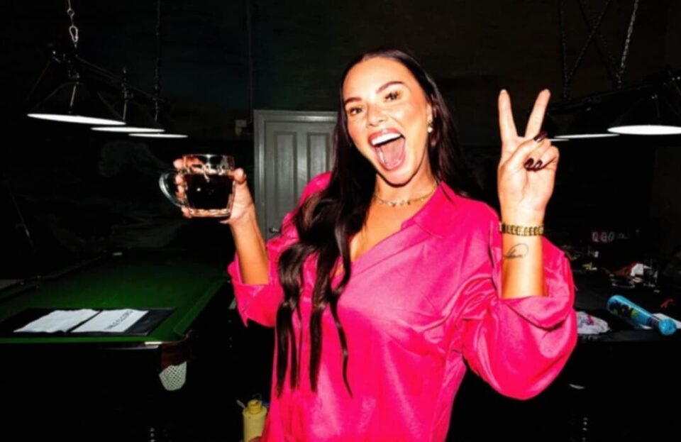 Rosie Clarke in a bright pink shirt holding a drink and flashing a peace sign, celebrating after a skimpy barmaid shift in WA.
