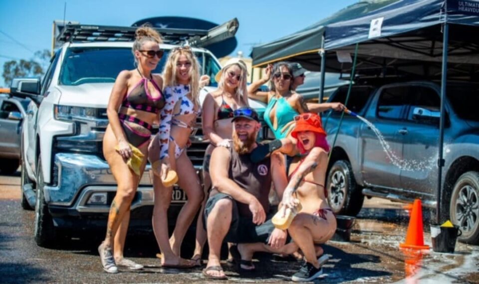 Group of smiling women in bikinis and a man pose together during a skimpy car wash event in WA, with sudsy vehicles and tents in the background.